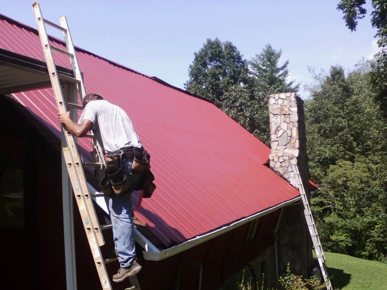 Roofer on a ladder beside a red metal roof and stone chimney — Soffit & Fascia Repair by Roofing Brothers in Una